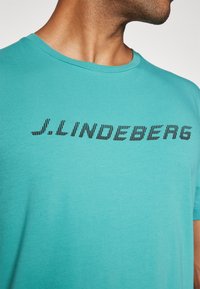 Turkio spalvos medvilnės marškinėliai su juodu horizontaliu „J. LINDEBERG“ logotipu ant krūtinės. Trumpi rankovės ir apvalus iškirptės. 