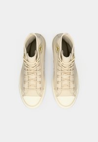 Beige kanvas høyde sneakers med gull logo, rund tå, gummisåle, og snøring; glatt tekstur med sømdetaljer.