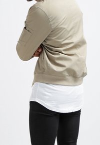 Beige bomberjacka med en slät textur, ribbad fåll och sidofickdetaljer. Lager ovanpå en lång vit skjorta och kombinerad med svarta byxor.