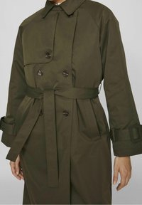 Oliwkowy trencz wykonany z gładkiego materiału, z podwójnym zapięciem z przodu, paskiem w talii oraz dużymi guzikami, z klasycznym kołnierzem.