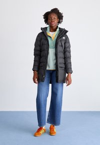 Svart pufferjakke med hette, glidelås foran og sidelommer, lagd over en teal polo-skjorte. Kombinert med vide blå jeans og oransje sneakers.
