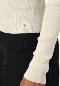 Detailní záběr na béžovou žebrovanou svetr s rukávy a lemem s nášivkou Calvin Klein Jeans, kombinovaný s černými kalhotami.