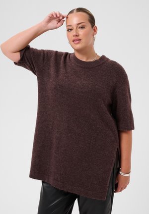 Femme portant un pull ample à manches courtes marron foncé et un pantalon noir, un bras levé près de sa tête, sur un fond uni.