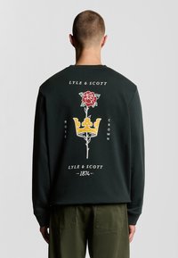 Dunkelgrüner Sweatshirt mit einem zentralen Grafikmotiv eines rosaroten Raben und einer Krone in Rot und Gelb, mit dem Text "Lyle & Scott" und "1874" darüber und darunter.