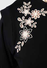 Zwarte stof met geborduurd bloemenmotief in lichtroze, met bloemen en bladeren in verschillende vormen en maten op het schoudergebied.