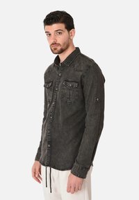 Camicia a maniche lunghe in denim nero con chiusura a bottoni, due tasche sul petto e cuciture a contrasto. Leggera trama usurata e cuciture delle spalle sagomate.