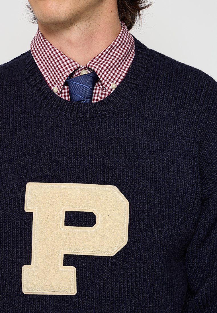 Marineblauer Strickpullover mit Rundhalsausschnitt und beigem Filz-"P"-Logo. Unterhalb sichtbar ist ein rot-weißes Karodresshemd, das mit einer blauen Krawatte kombiniert ist.