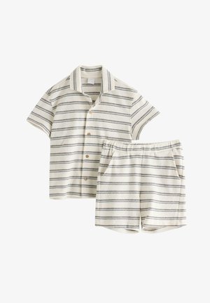 Junger Junge trägt beige Kappe, gestreiftes kurzärmeliges Hemd und passende Shorts, steht mit einer Hand in der Tasche und braunen Sandalen.