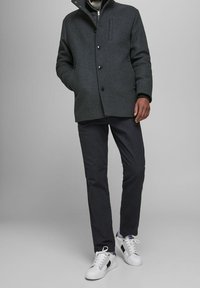 Cappotto di lana grigio con colletto alto e chiusura a bottoni frontali, abbinato a pantaloni scuri e scarpe da ginnastica bianche con accenti di colore discreti.