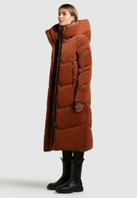 Cappotto lungo e voluminoso di colore ruggine, con grande colletto e chiusura con zip, caratterizzato da cuciture orizzontali e tasche laterali, indossato con stivali neri.
