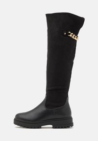 Las botas negras hasta la rodilla presentan una mezcla de texturas de cuero y ante, con un acabado suave en la sección inferior y un acento de cadena dorada en el lateral.