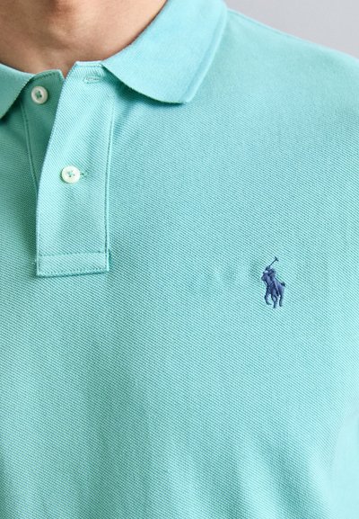 Ljusblå polo skjorta i texturerad tyg, med en tre-knapps halsringning och en broderad mörkblå logotyp på vänster bröst.