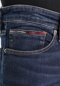 Mörkblå denimjeans med ett slitet fickkant, med subtila röd- och vitstickningar och en silverfärgad knappdetalj.