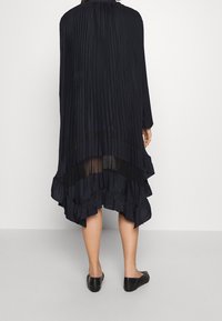 Černé skládání šaty s dlouhými rukávy a volánkovým lemem. Průhledná vrstva viditelná vzadu. Nosené ploché černé texturované boty.