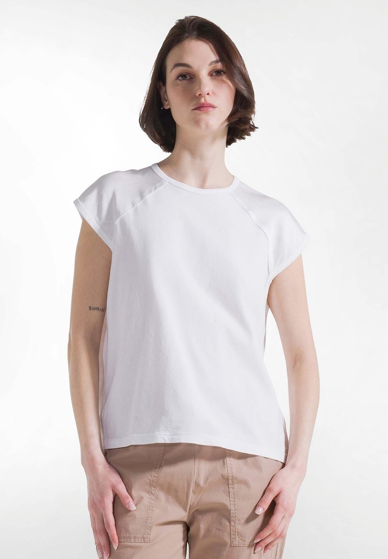 Femme aux cheveux bruns courts, portant une chemise blanche à manches courtes et un pantalon beige, debout devant un fond blanc uni.