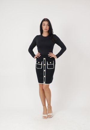 Frau steht mit den Händen in der Hüfte, trägt ein schwarzes langärmliges Oberteil und einen schwarzen Rock mit weißer Einfassung und goldenen Knöpfen, weiße Sandalen.