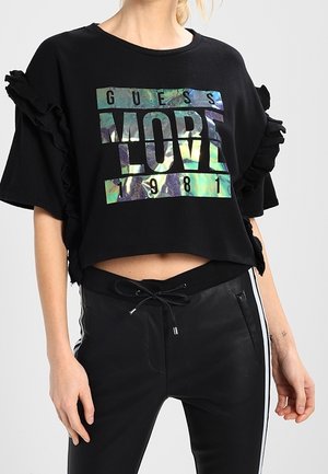 Nainen pukeutunut mustaan lyhennettyyn t-paitaan, jossa on helmiäishohtoinen teksti "GUESS MORE LOVE 1981" ja röyhelöitä, yhdistettynä mustiin nyörivöisiin housuihin.