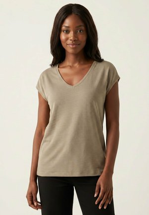 Femme aux longs cheveux foncés portant un haut beige à manches courtes et col en V et un pantalon noir, debout devant un fond clair uni.