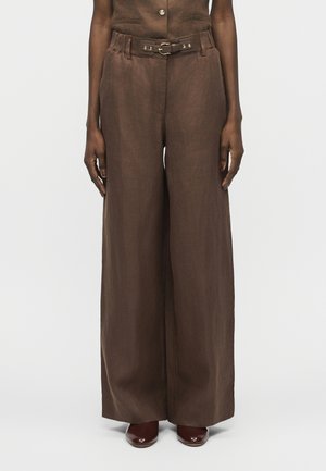 Καφέ παντελόνια με φαρδιά μπατζάκια και ψηλή μέση, με ζώνη στο ίδιο χρώμα, φορεμένα με σκούρα καφέ παπούτσια σε ένα άτομο που στέκεται όρθιο.