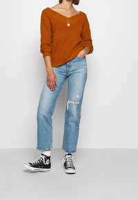 Pullover in Rostrot mit weitem Ausschnitt, kombiniert mit hellblauen, ausgewaschenen Jeans mit geradem Bein und schwarzen High-Top-Sneakers.