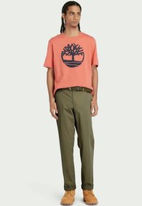Orange cotton t-shirt with tree graphic, paired with olive green trousers. Tan footwear completes the look, emphasizing a casual style.