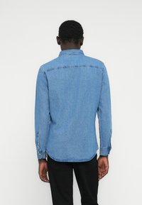 Világoskék farmering gallérral, hosszú ujjakkal és gombos mandzsettával. Ívelt szegéllyel és látható varrási részletekkel.