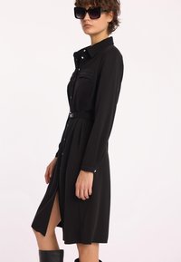 Czarna sukienka koszulowa z długimi rękawami, z guzikami z przodu, dwoma kieszeniami na piersi, paskiem w talii i rozcięciem z boku. Noszona z dużymi okularami przeciwsłonecznymi.