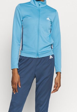 Osoba nosząca jasnoniebieską kurtkę Adidas rozpinaną na zamek i ciemnoniebieskie spodnie Adidas z białymi paskami na jednolitym tle.