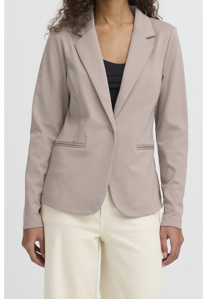 Vrouw die een beige, nauwsluitend colbert draagt met een enkele knoop, gecombineerd met een zwarte top en crèmekleurige broek, staand met ontspannen armen.