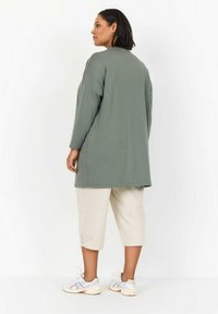 Tunic a maniche lunghe, verde oliva, con una vestibilità comoda, abbinata a pantaloni beige a 3/4 con dettagli di bottoni, indossati con scarpe da ginnastica bianche.