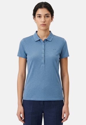 Donna in piedi, di fronte, che indossa una polo a maniche corte azzurro chiaro e pantaloni blu scuro su sfondo neutro.