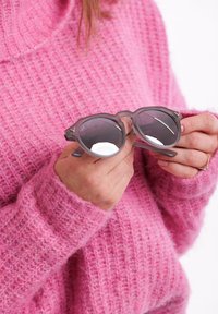 Šedé sluneční brýle s kulatými obroučkami a tmavými skly držené v rukou, položené na texturovaném růžovém pleteném svetru.