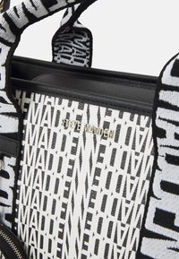 Gros plan d'un sac à dos Steve Madden noir et blanc avec un motif répétitif du nom de la marque et des poignées en tissu rembourré.