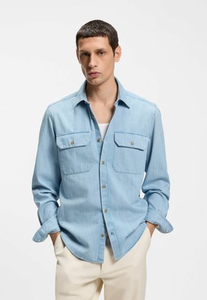 Giovane uomo con capelli corti e scuri che indossa una camicia di jeans azzurro chiaro sopra una maglietta bianca e pantaloni beige, con le mani nelle tasche, in piedi contro uno sfondo bianco.