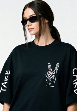 Frau mit dunklem Haar im Pferdeschwanz, die schwarze Sonnenbrille und ein schwarzes T-Shirt mit dem Peace-Handzeichen und den Worten Glaube, Liebe, Hoffnung, Frieden trägt.