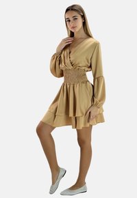 Abito avvolgente beige in satin con maniche lunghe, vita arricciata e gonna a strati. La modella indossa ballerine argentate scintillanti. Design semplice ed elegante.