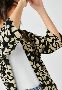 Locker sitzender Kimono mit einem abstrakten geometrischen Muster in Schwarz und Beige, aus leichtem Stoff gefertigt. Verfügt über weite Ärmel und eine offene Front.