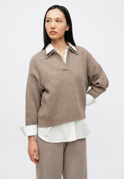 Naine, kes kannab beeži kootud kampsunit ja pükse valge särgiga all, seisab ühe käega puusal, vaadates kõrvale.