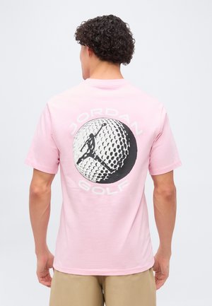 Mężczyzna w różowym T-shircie z dużym wzorem piłki golfowej i logo Jumpman oraz napisem „Jordan Golf” na plecach, widoczne beżowe szorty poniżej.