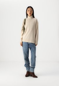 Pull à col roulé beige en tissu doux, associé à un jean bleu ample et des bottes à talons brunes avec des détails complexes.