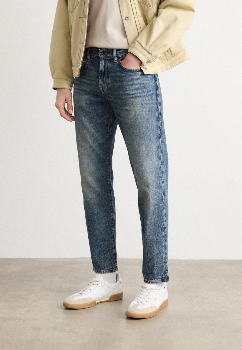 Jeans din denim de un albastru deschis, cu o croială dreaptă, având accente decolorate și buzunare standard. Asortate cu sneakers albi și o jachetă bej.