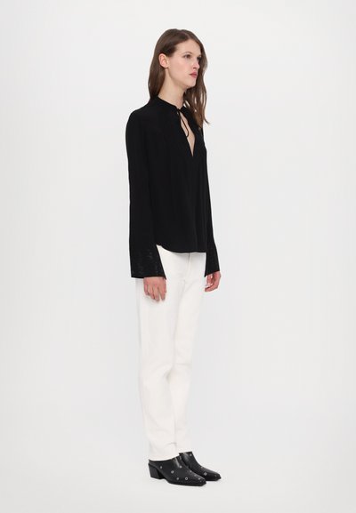 Black long-sleeve blouse with a V-neckline, featuring textured detailing at the cuffs, paired with white trousers and black heeled shoes.