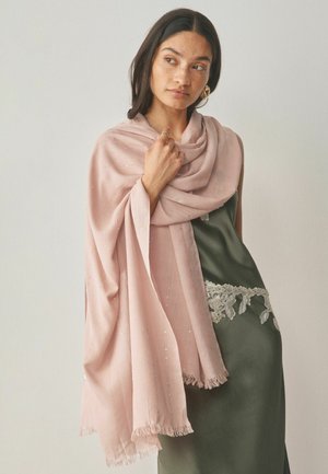 Woman with dark hair wearing gold earrings and a green lace-trimmed dress draped in a soft pink shawl with fringe edges.