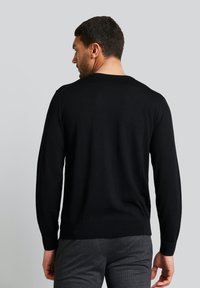 Schwarzer Pullover mit langen Ärmeln, runden Ausschnitt, gerippten Bündchen und Saum, weiche Textur. Modell steht mit dem Rücken zur Kamera und trägt grau gemusterte Hosen.