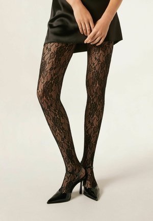 Persona que lleva mallas de encaje floral negro combinadas con unos tacones de punta negra de charol y un vestido de satén negro.