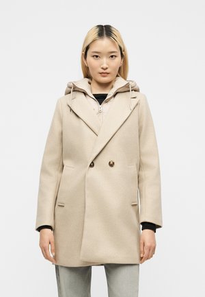 Jeune femme portant un manteau en laine beige croisé sur un sweat à capuche zippé beige avec un pantalon gris clair, debout devant un fond blanc uni.