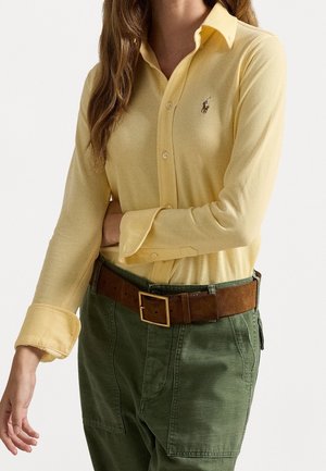 Kobieta ubrana w żółtą koszulę na guziki z długim rękawem i małym logo, włożoną do zielonych spodni z brązowym paskiem, na białym tle.