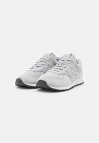 Vaaleanharmaat New Balance -kengät, jotka on valmistettu mokasta ja joissa on kangaskoristeluja. Kenkäparissa on teksturoitu kumipohja, nyöritysdesign ja logo sivussa.