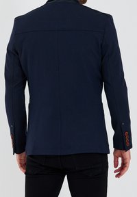 Mörkblå jacka med texturerad tyg, med en slät krage och knapputskrifter vid manschetten. Skräddarsydd design med rak rygg.