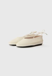 Chaussures plates beiges à bout rond, au design minimaliste, avec de fines brides à la cheville. La semelle est noire avec une texture lisse.
